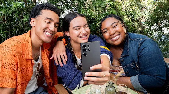 Selfie için gülümseyen üç kişi arasından ortadaki kişi, Android uyumlu telefonuyla bu anı yakalıyor.
