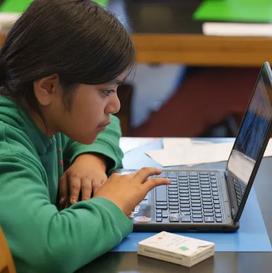 A child with long, black hair in a ponytail, wearing a green hooded sweatshirt, intently uses a laptop on a table.