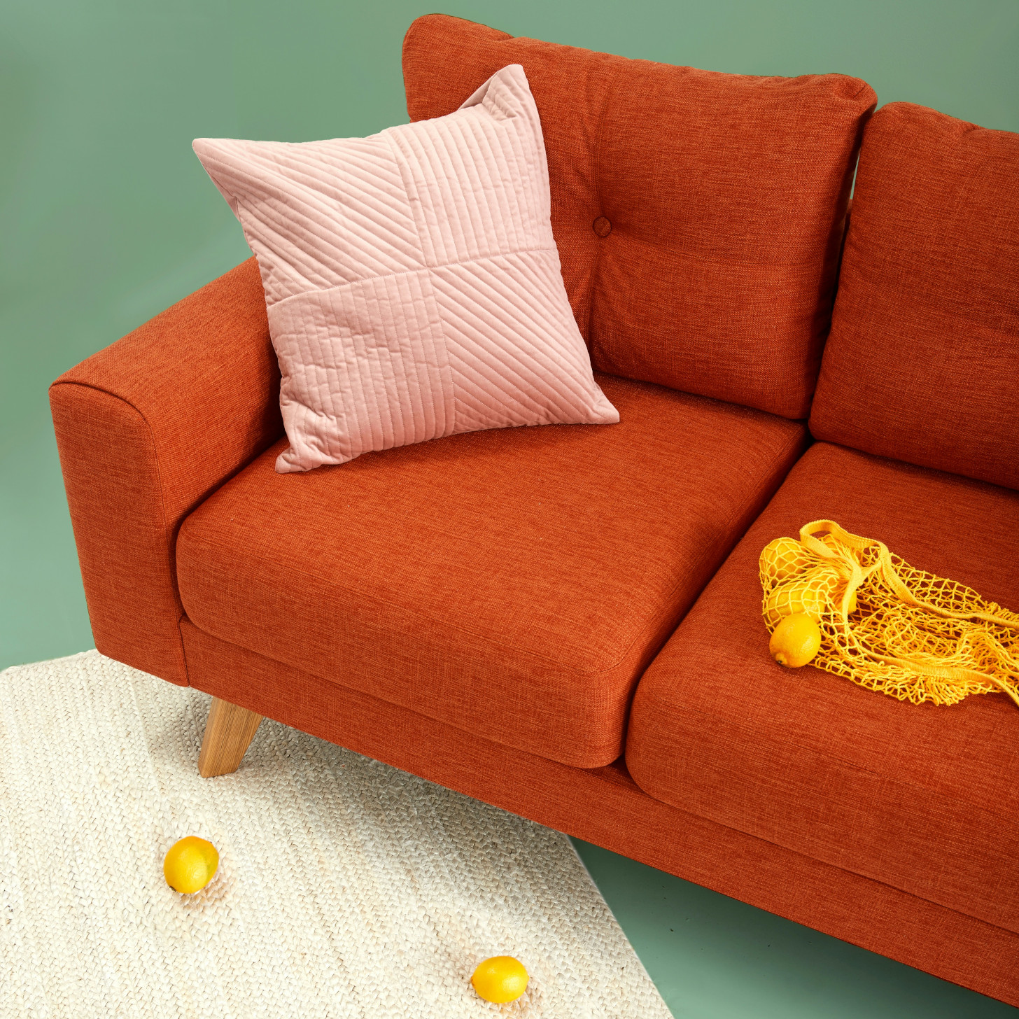 Sample base image of a red couch that has a bag of lemons placed
           on the right and a throw pillow on the left corner. The couch is
           placed at an angle, with a single foot placed on a white area rug. In
           front of the couch on the area rug are two lemons.