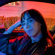A person sitting in a car at night. Their face appears in sharp detail despite the scene's low red and blue light.